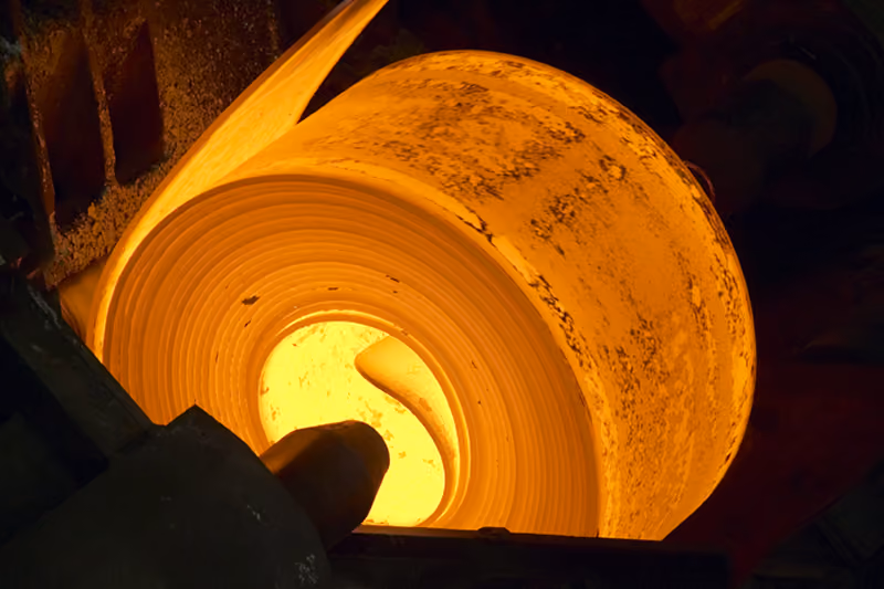 A glowing hot coil of metal being formed or processed in an industrial setting, emitting bright orange and yellow light.