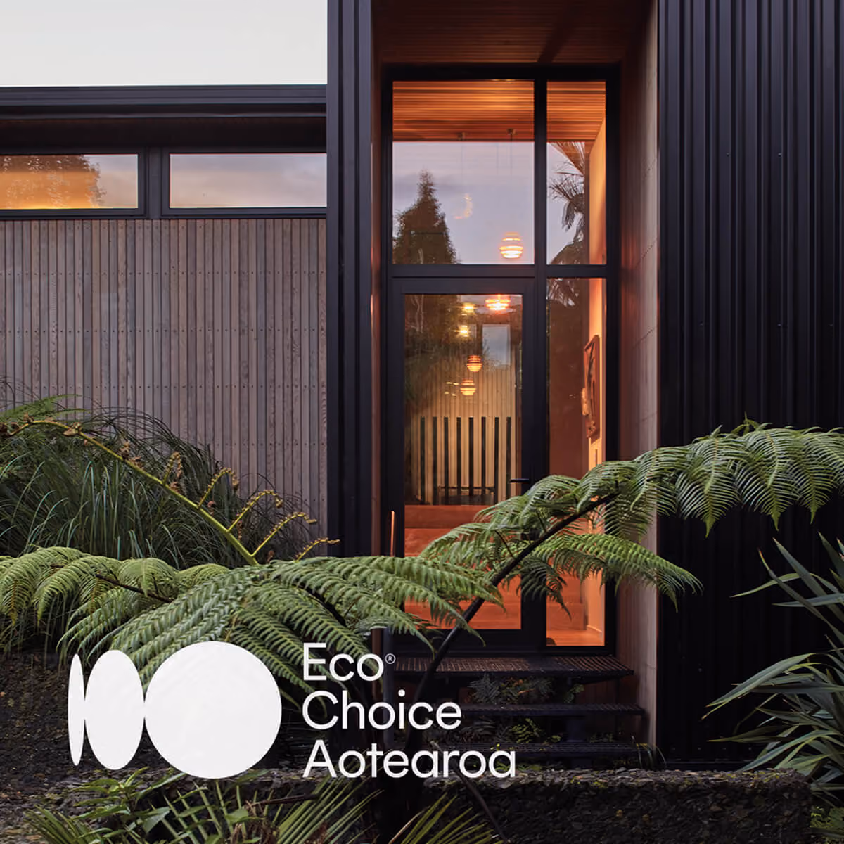 Modern building entrance with large glass door framed by black panels and wooden siding, surrounded by lush green ferns and plants, with warm indoor lighting visible through the door.