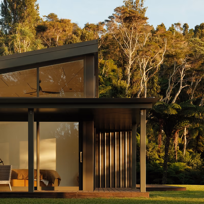 Modern house with large glass windows and a shaded porch, surrounded by lush green trees and lawn in golden sunlight.