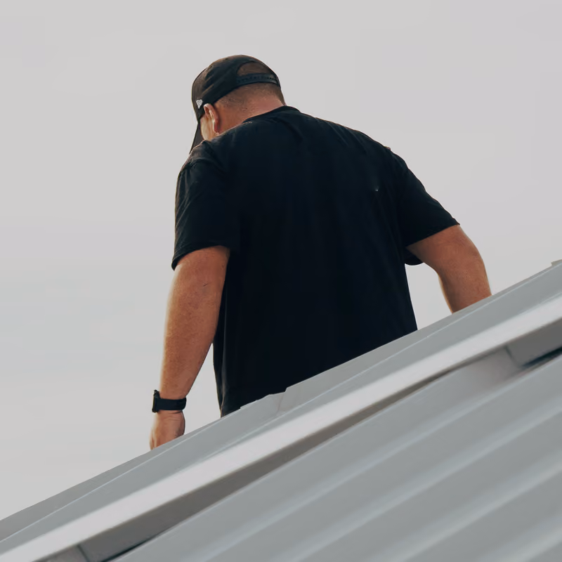 Man wearing a black t-shirt, black cap, and wristwatch standing on a metallic roof, viewed from behind against a pale sky.