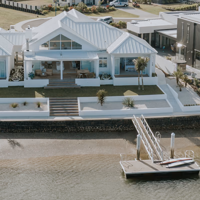 Modern white coastal house with metal roof, large windows, outdoor seating on wooden deck, landscaped garden, and private dock extending into calm water.