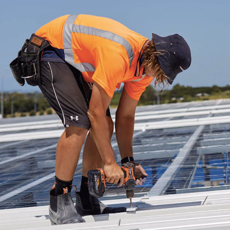 Worker wearing an orange high-visibility shirt and black shorts using a power drill to install solar panels on a rooftop under a clear sky.