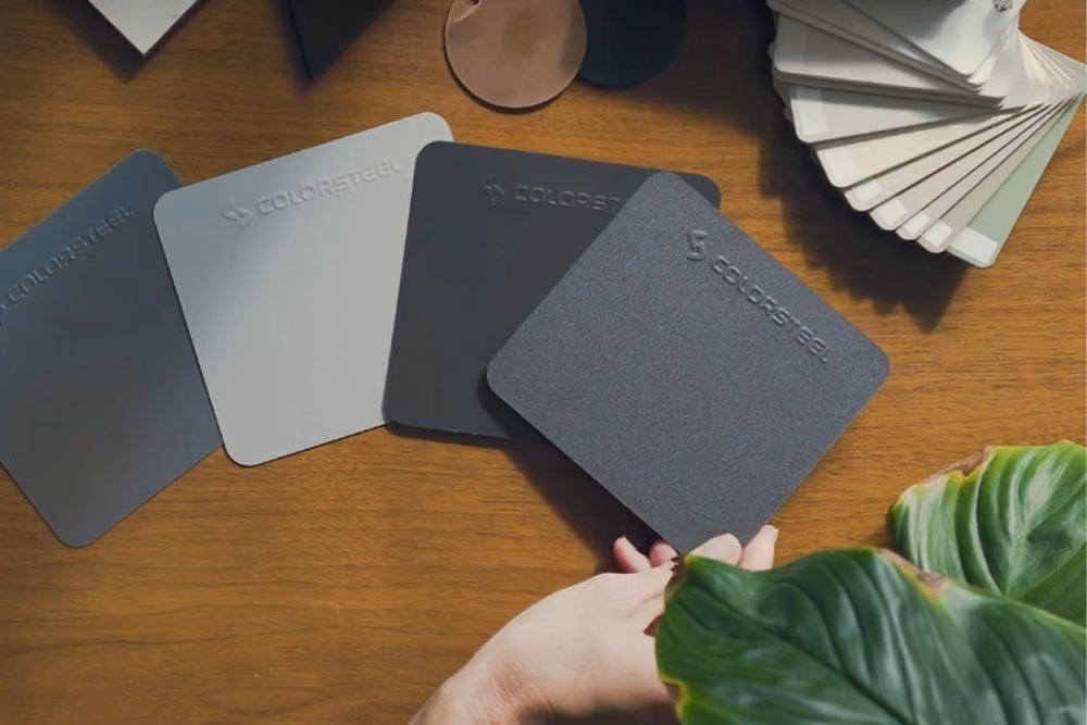 A hand holds a square dark grey Colorsteel sample on a wooden table next to three other Colorsteel samples in varying grey tones and a fan of white and grey color swatches, with a large green leaf in the foreground.