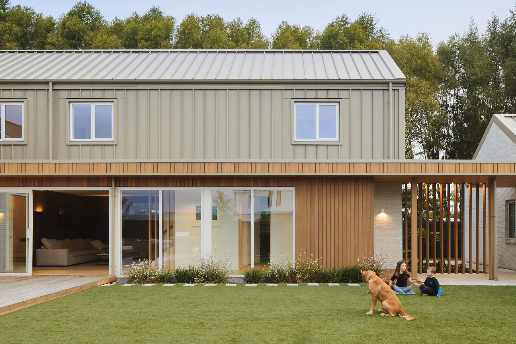 Modern two-story house with a green exterior and large glass sliding doors opening to a well-maintained lawn, where two children sit on the grass and a golden retriever dog faces them near the right side of the yard, surrounded by trees in the background.