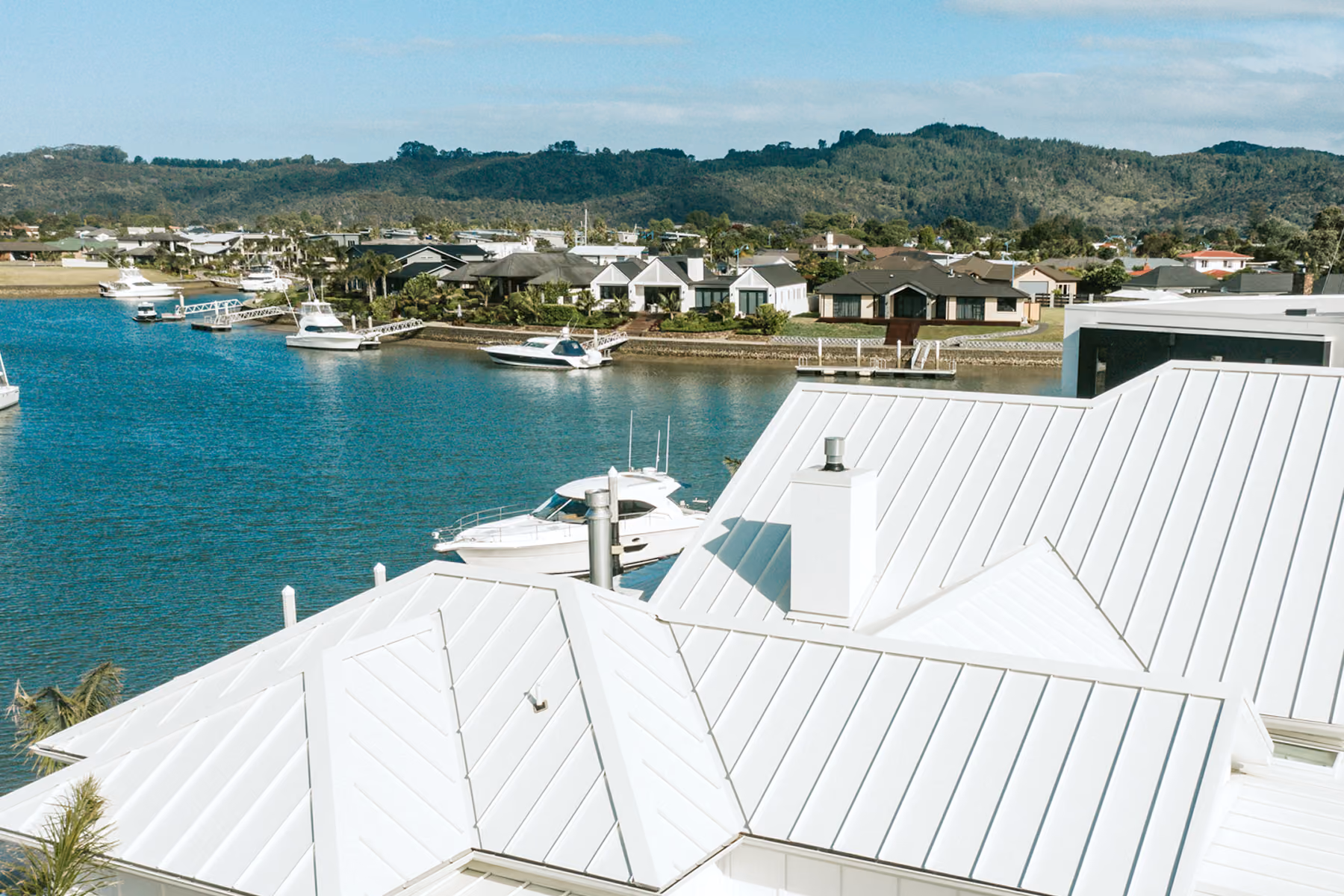 View of a waterfront neighbourhood with white metal-roofed houses in the foreground, boats moored on calm blue water, and wooded hills under a partly cloudy sky in the background.