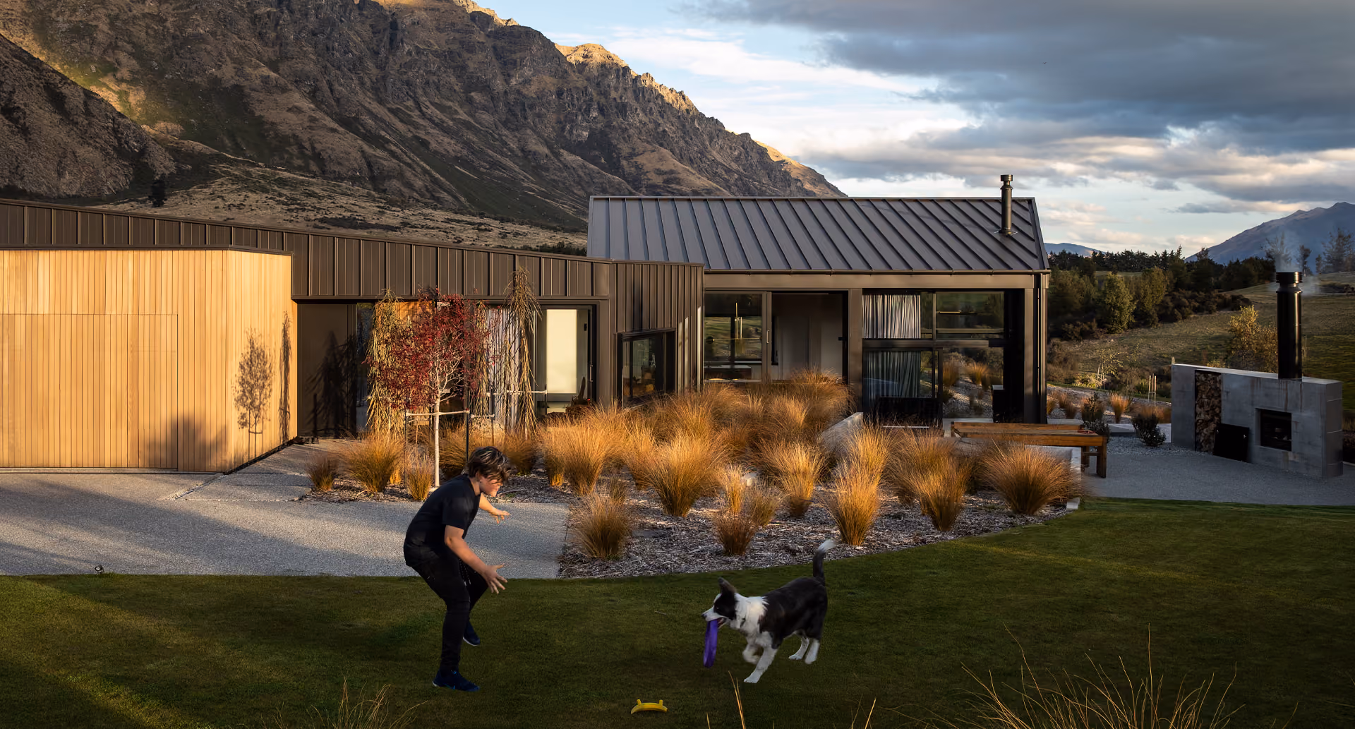 Man playing fetch with a black and white dog on a green lawn outside a modern house with brown and grey exterior, surrounded by mountain landscape under a partly cloudy sky.
