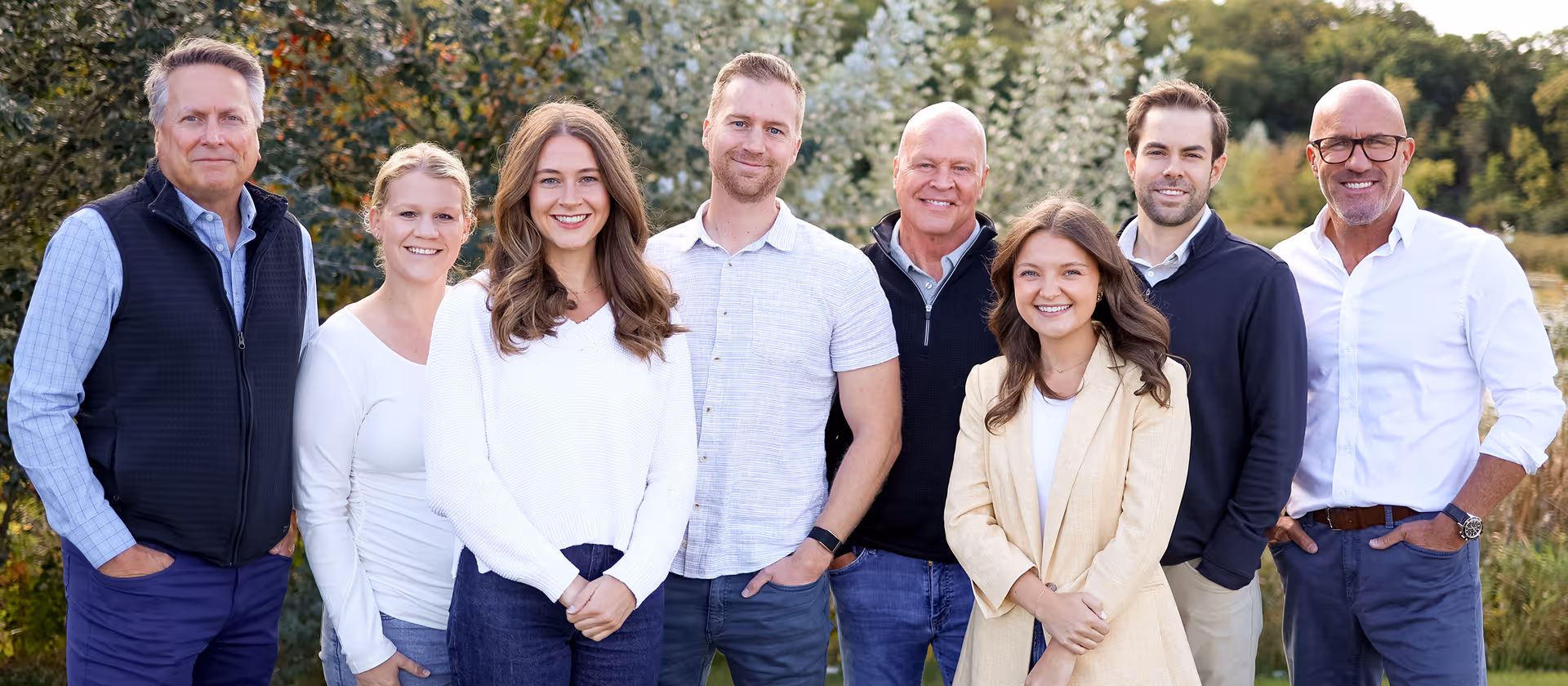 Group portrait of eight smiling people standing outdoors in casual and business casual attire.