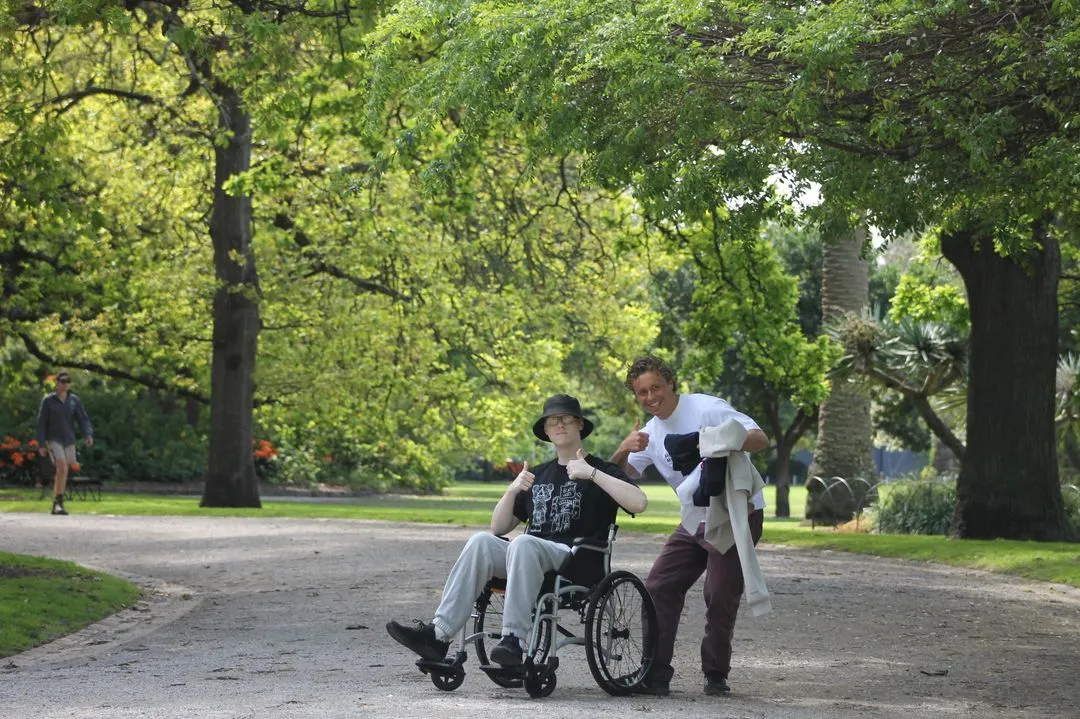 Two young men giving thumbs up; one in a wheelchair, the other standing beside him in a park.