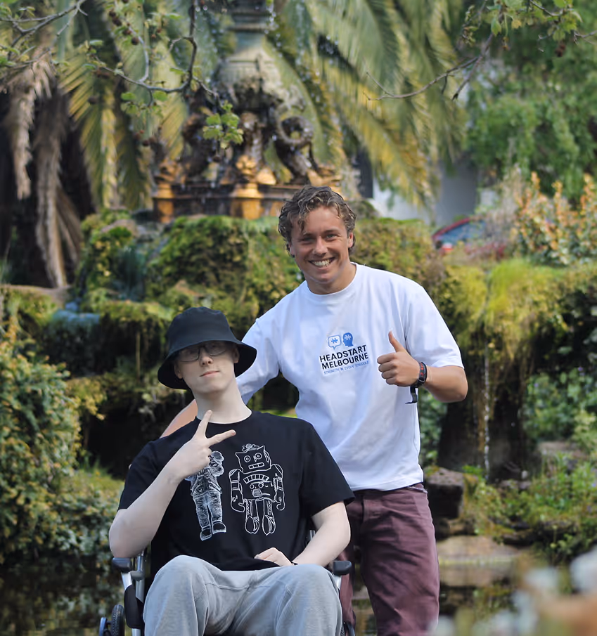 Smiling young man giving thumbs up behind a person in a wheelchair posing with peace sign outdoors.
