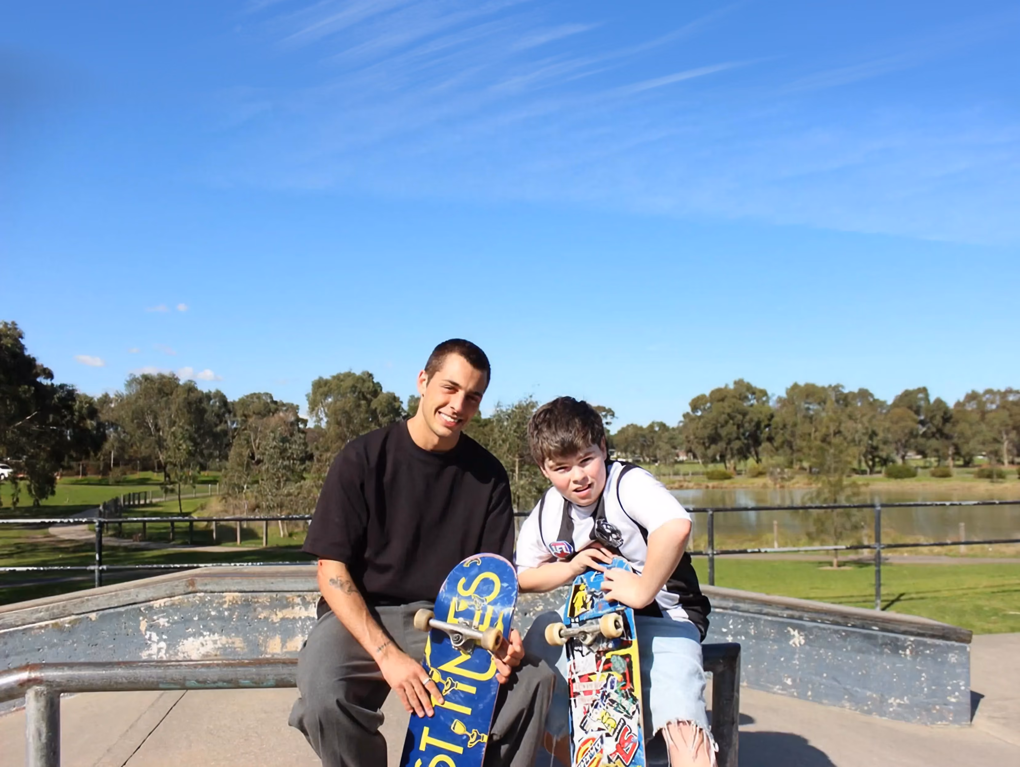 Man and boy sitting on skateboards at skate park with trees and lake in background.