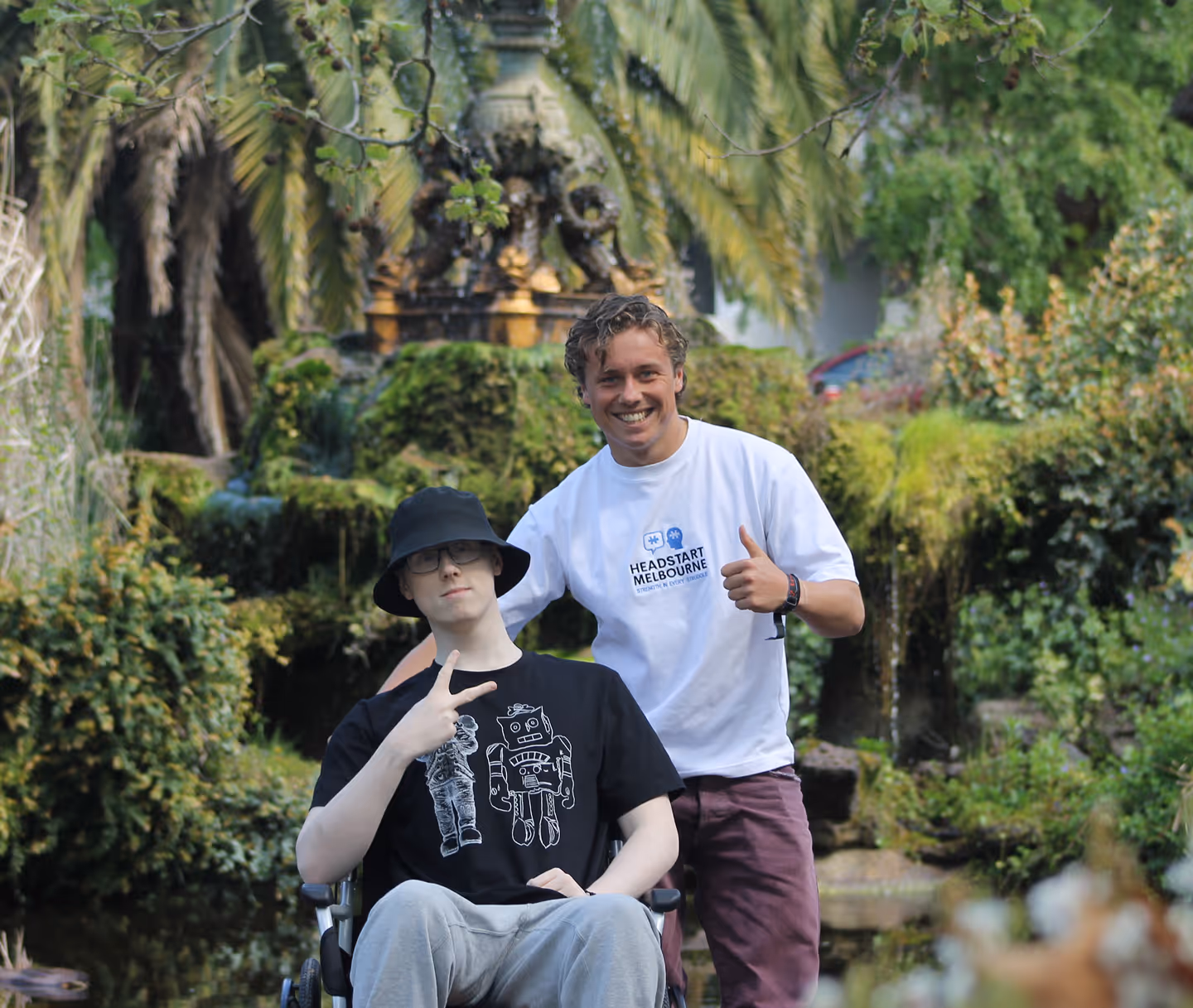Two young men outdoors, one in wheelchair showing peace sign, the other giving thumbs up.