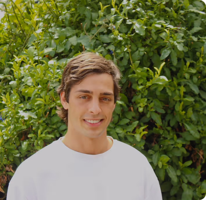 Young man with brown hair wearing a white shirt standing in front of leafy green bushes outdoors.
