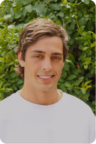 Smiling young man with curly hair and mustache wearing a white shirt, standing before purple flowers.