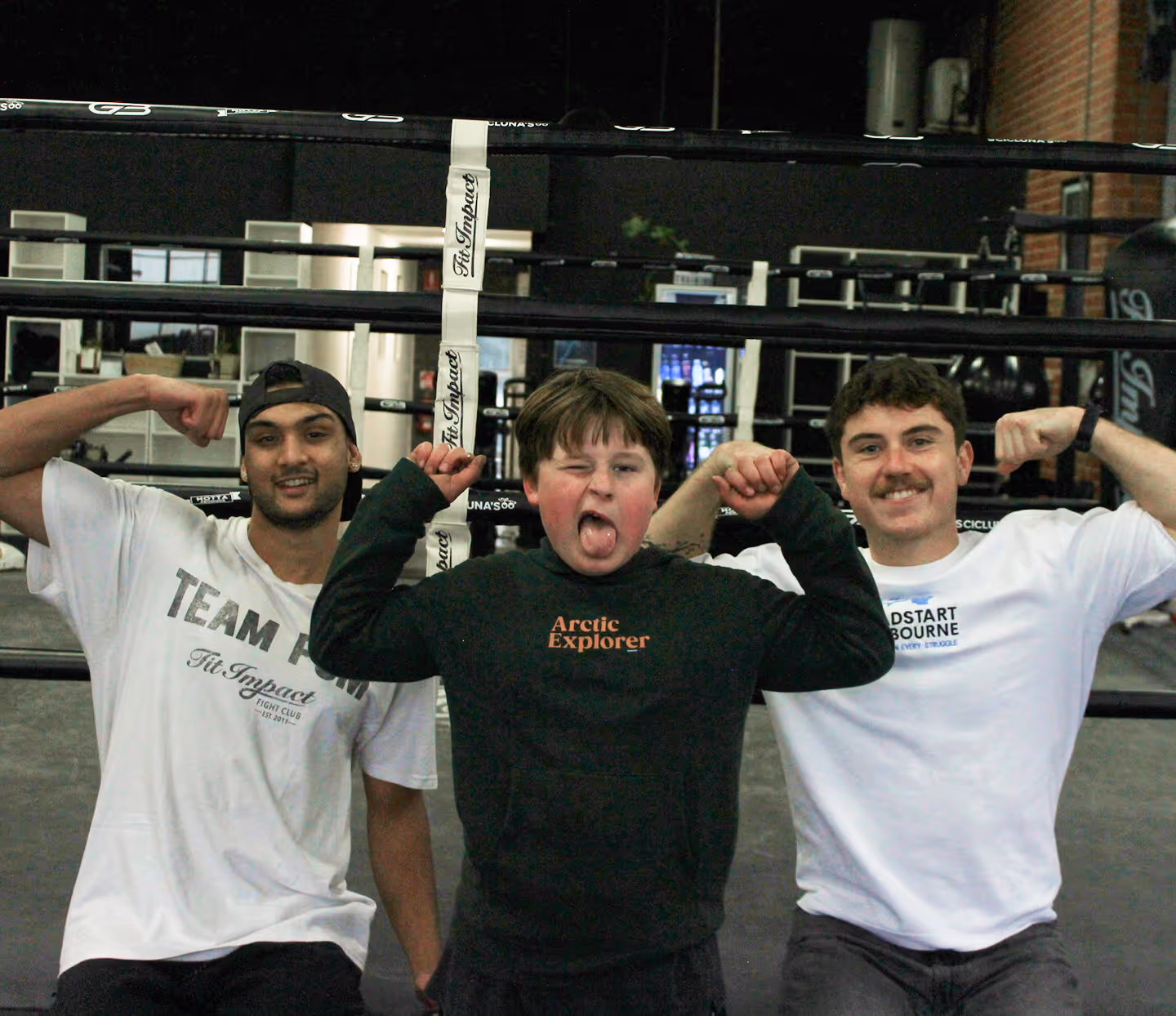 Three people in a boxing gym flexing arms; middle person winks and sticks out tongue.