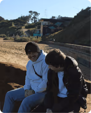 Two young men laughing and talking in a sunlit alleyway with brick walls and greenery.