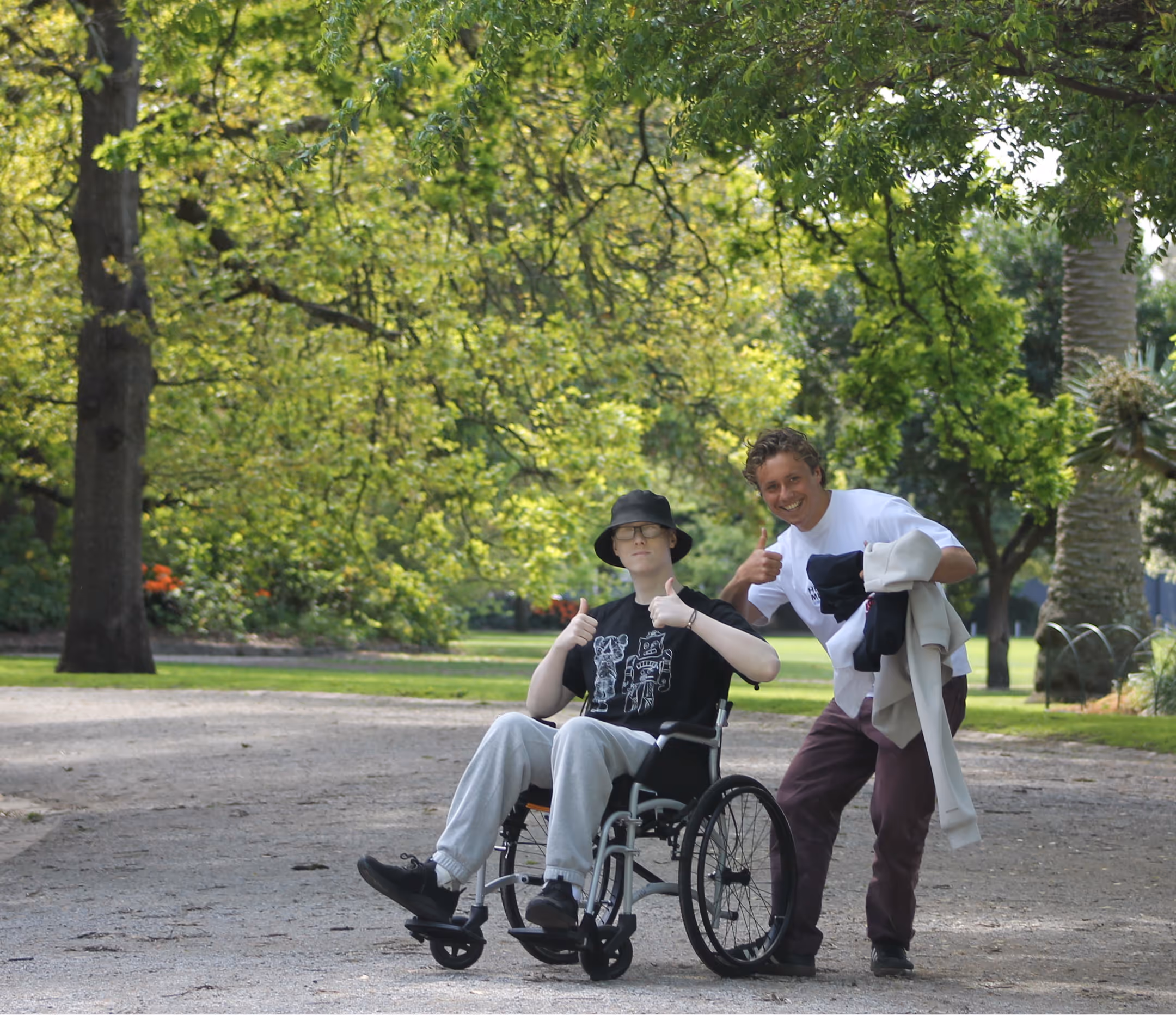 Person in wheelchair and another person beside them outdoors, both giving thumbs up in a park.