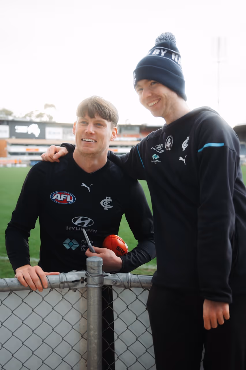 Two men in Carlton Football Club gear smiling by a fence on a football field.