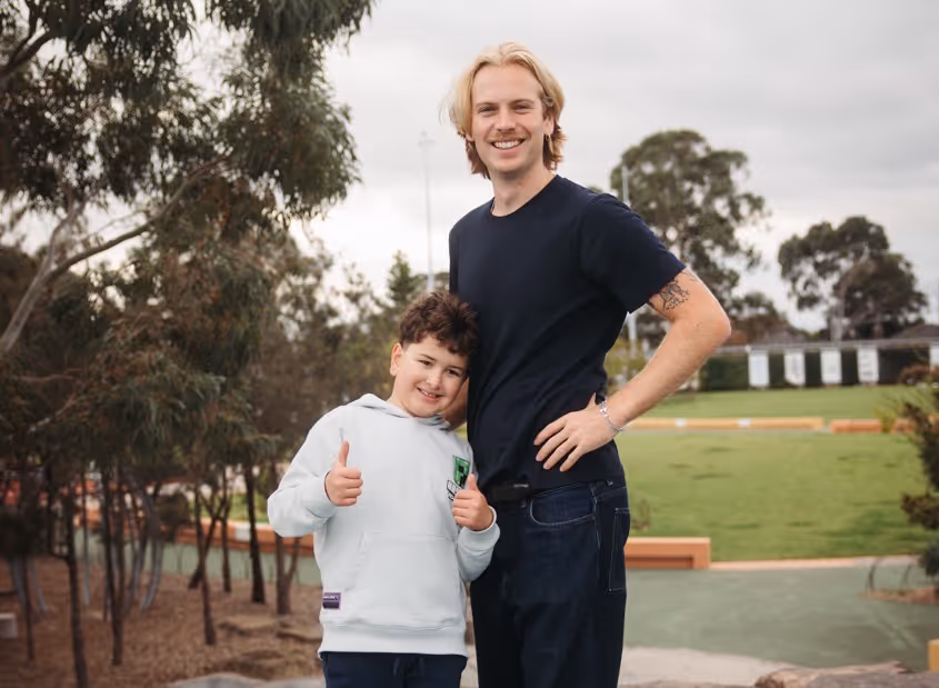 Smiling young man with a tattoo stands next to a boy giving thumbs up in an outdoor park.