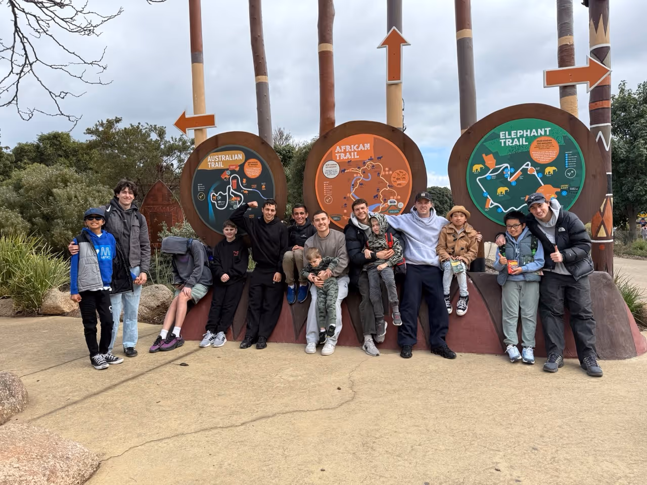 Group of people posing in front of Australian, African, and Elephant Trail signs in outdoor park.