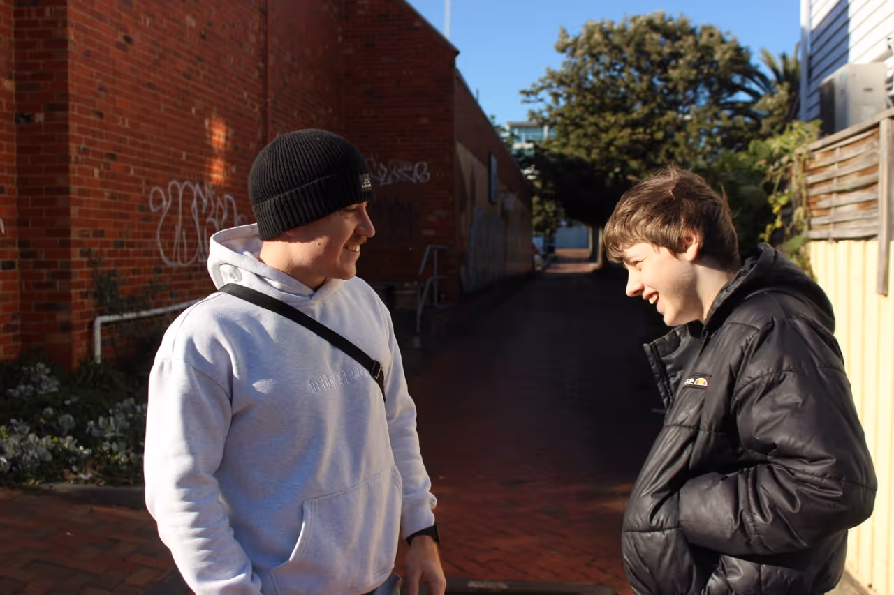 Two young men smiling and talking in an alley with brick walls and trees in sunlight.