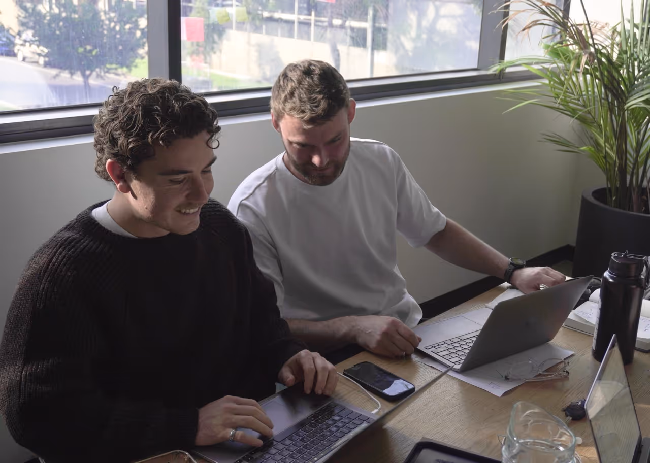 Two men sitting at a desk working on laptops near a window and a potted plant.