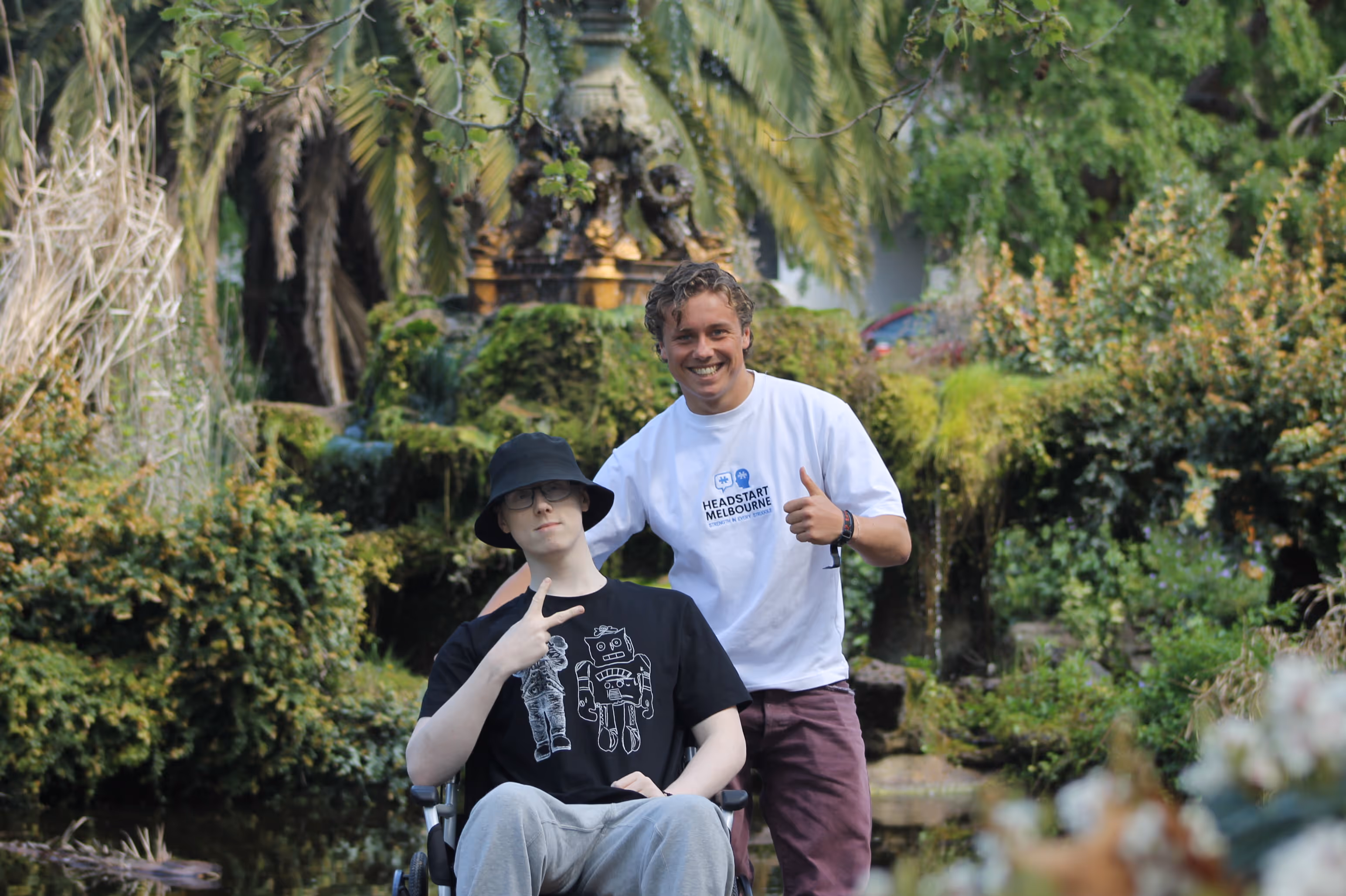 Two young men smiling in a garden, one sitting in a wheelchair making a peace sign.