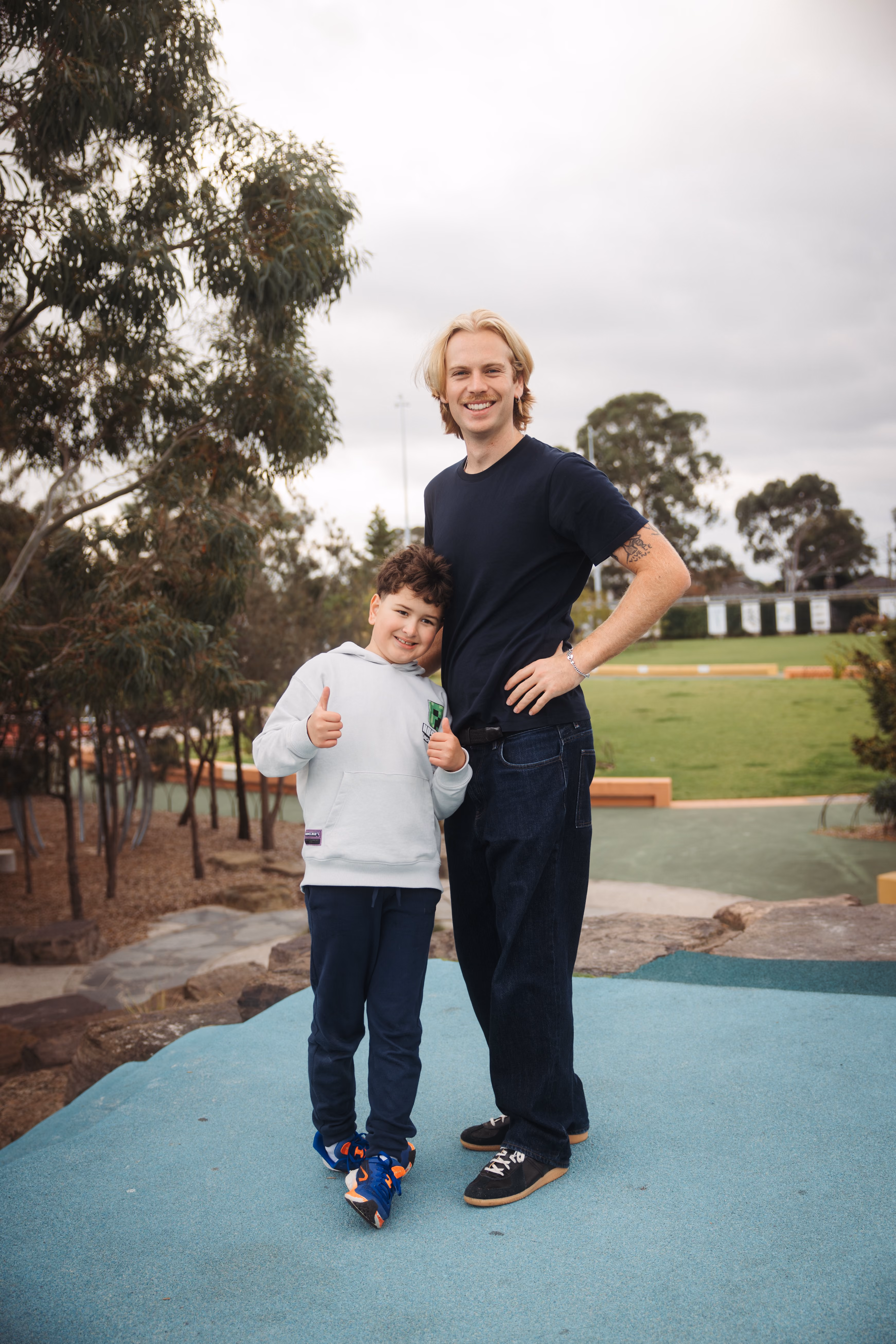 Smiling man with blond hair stands beside boy in white hoodie giving thumbs up outdoors.
