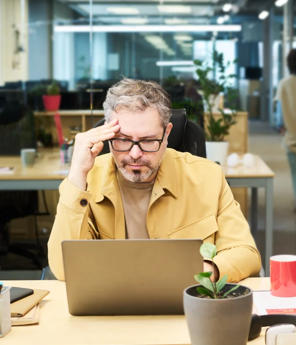 Man wearing glasses and tan jacket working on a laptop in a modern office with plants.
