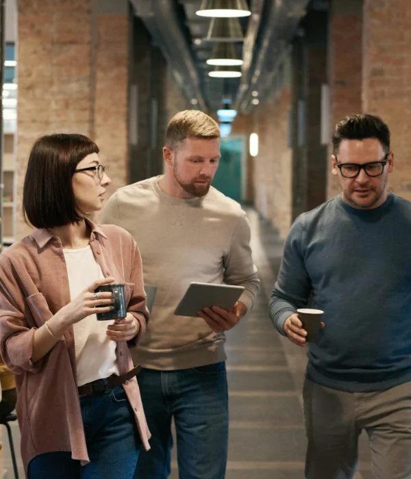 Three coworkers walking in an office hallway discussing work, holding a tablet and coffee cups.