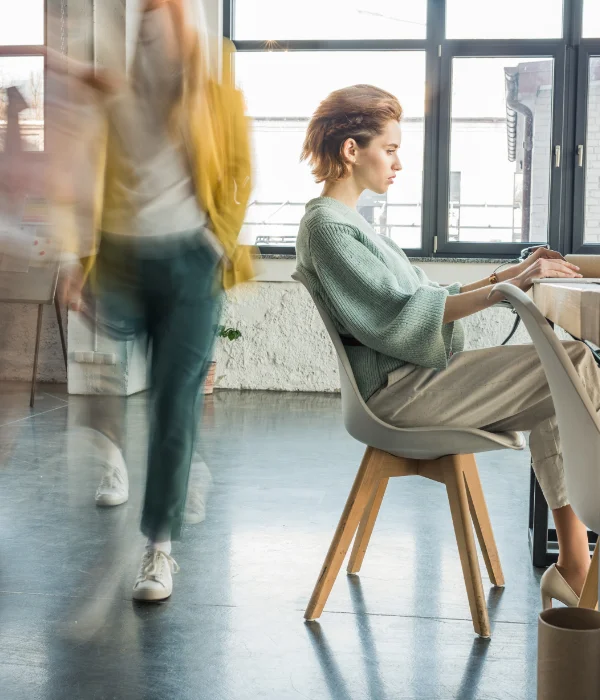 Focused woman in green sweater typing at desk with blurred figures walking nearby in modern office.