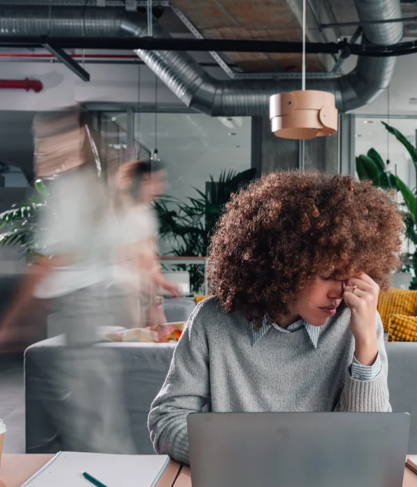 Tired woman with curly hair resting her face on her hand working on laptop in busy office.