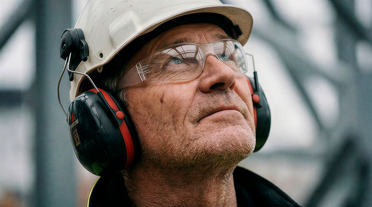 Close-up of male construction worker wearing white hard hat, safety glasses, and ear protection looking upward.