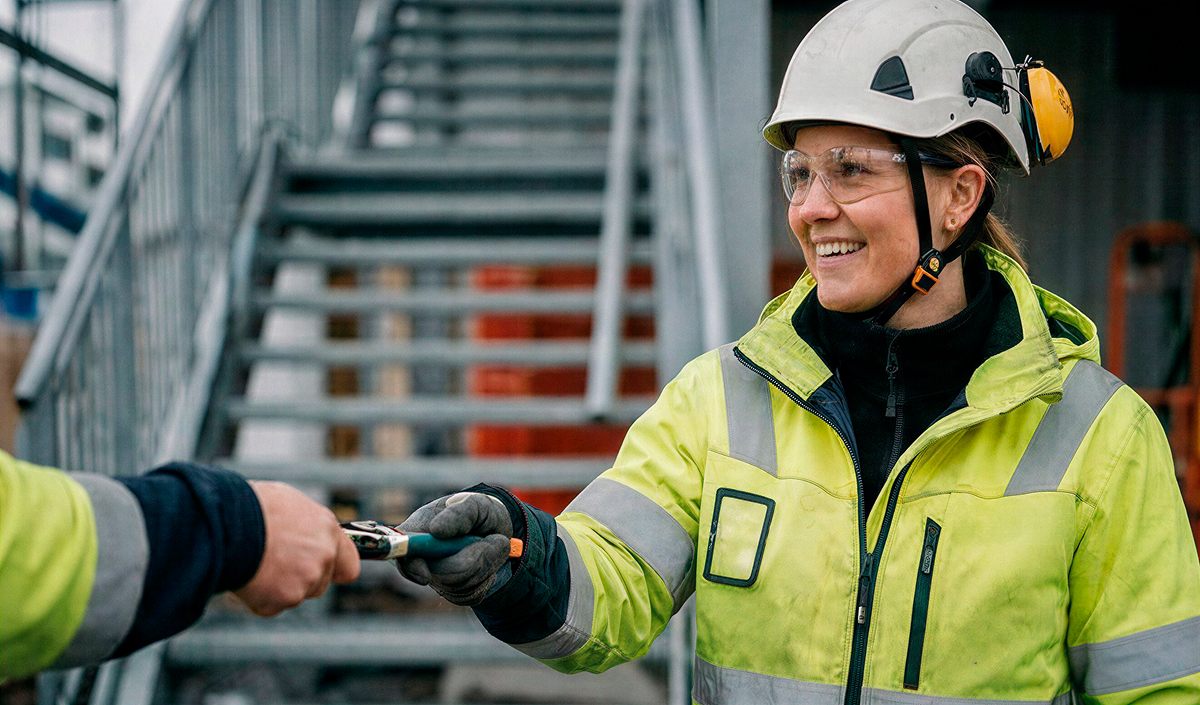 Worker in safety gear smiling and handing a tool to a colleague on an industrial site with stairs.