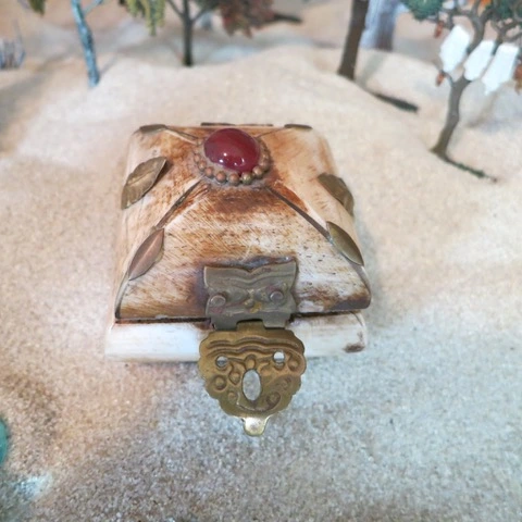 Small decorative box with a red stone and metal leaf designs on a sandy surface.