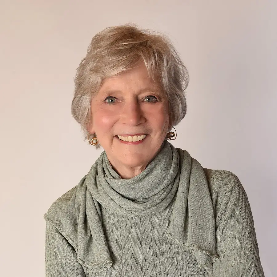 Smiling Dr. Hartshorn with short gray hair wearing a sage green sweater and scarf with gold earrings.