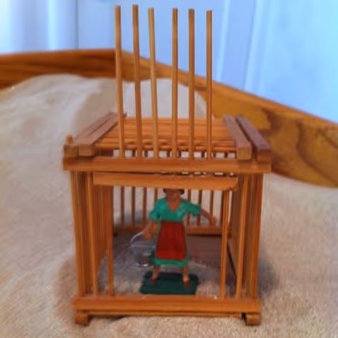 Miniature figure of a woman in a cage made of wooden sticks on a beige surface.