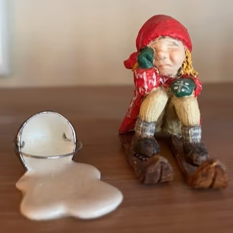 Small figurine of a skier wiping a tear while sitting on skis, next to a melting snowman ring holder.