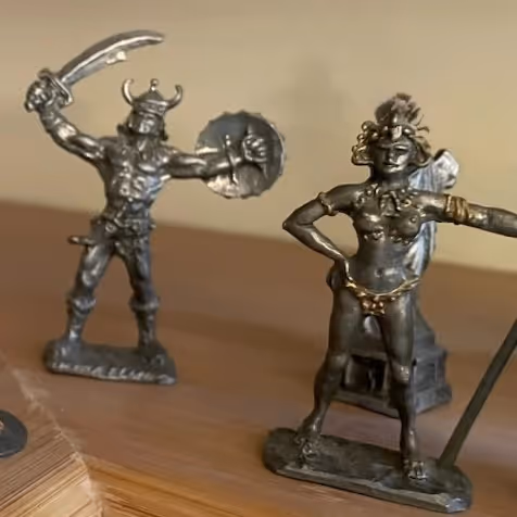 Two metal warrior figurines on a wooden surface, one holding a sword and shield, the other with hands on hips.