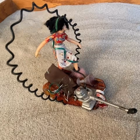 Miniature doll in floral dress sitting on a small mechanical vehicle with a coiled wire frame around it.