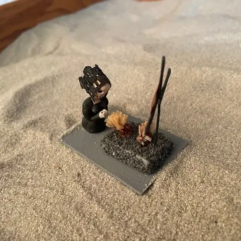 Miniature figure kneeling beside a small grave with flowers and tall sticks on sand.