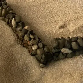 Corner of a small wall made with gray and black stones on beige sand.