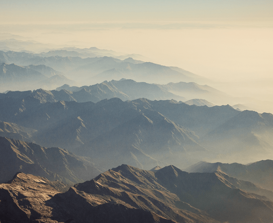 Layered mountain ranges fading into mist under a hazy sky at sunrise or sunset.
