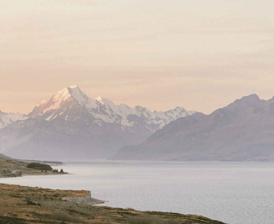 Snow-capped mountain range reflecting soft light above a calm lake with grassy shoreline.