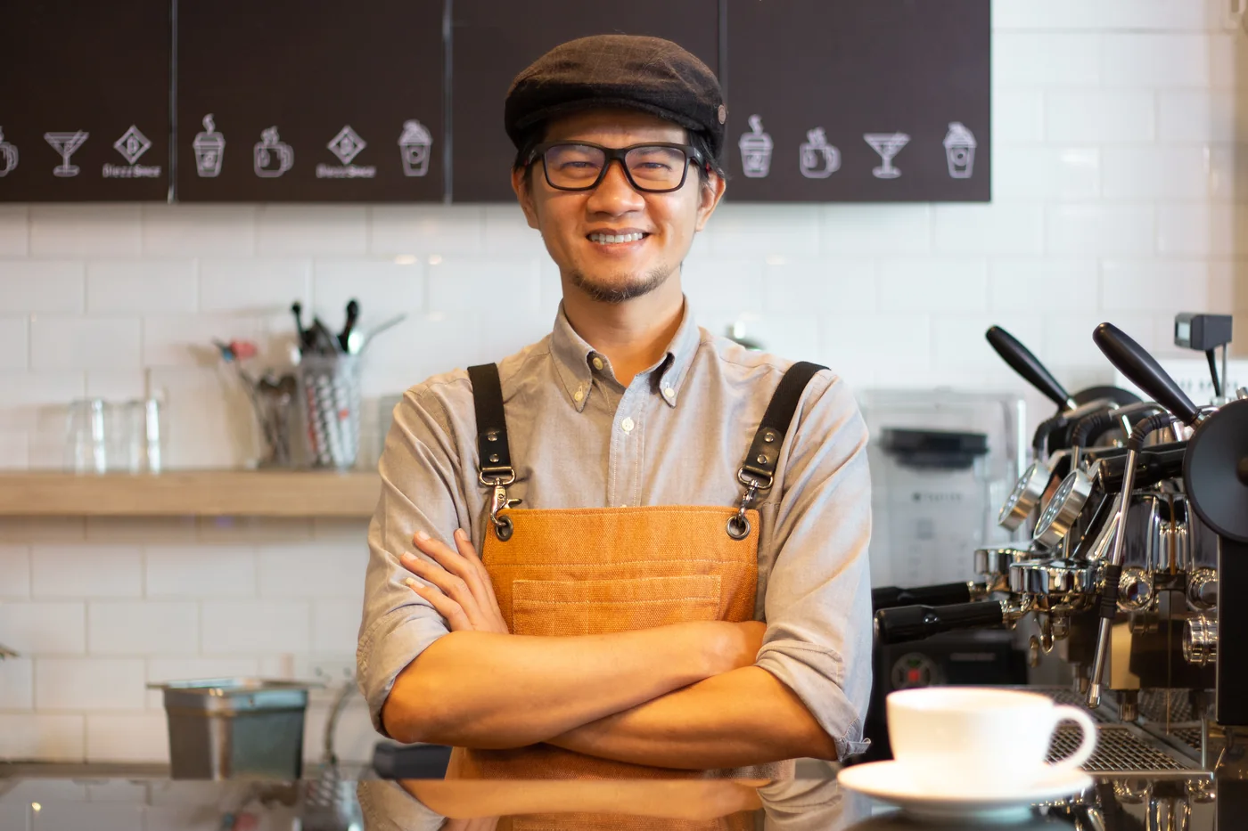Barista in an apron standing with arms crossed behind the coffee bar.