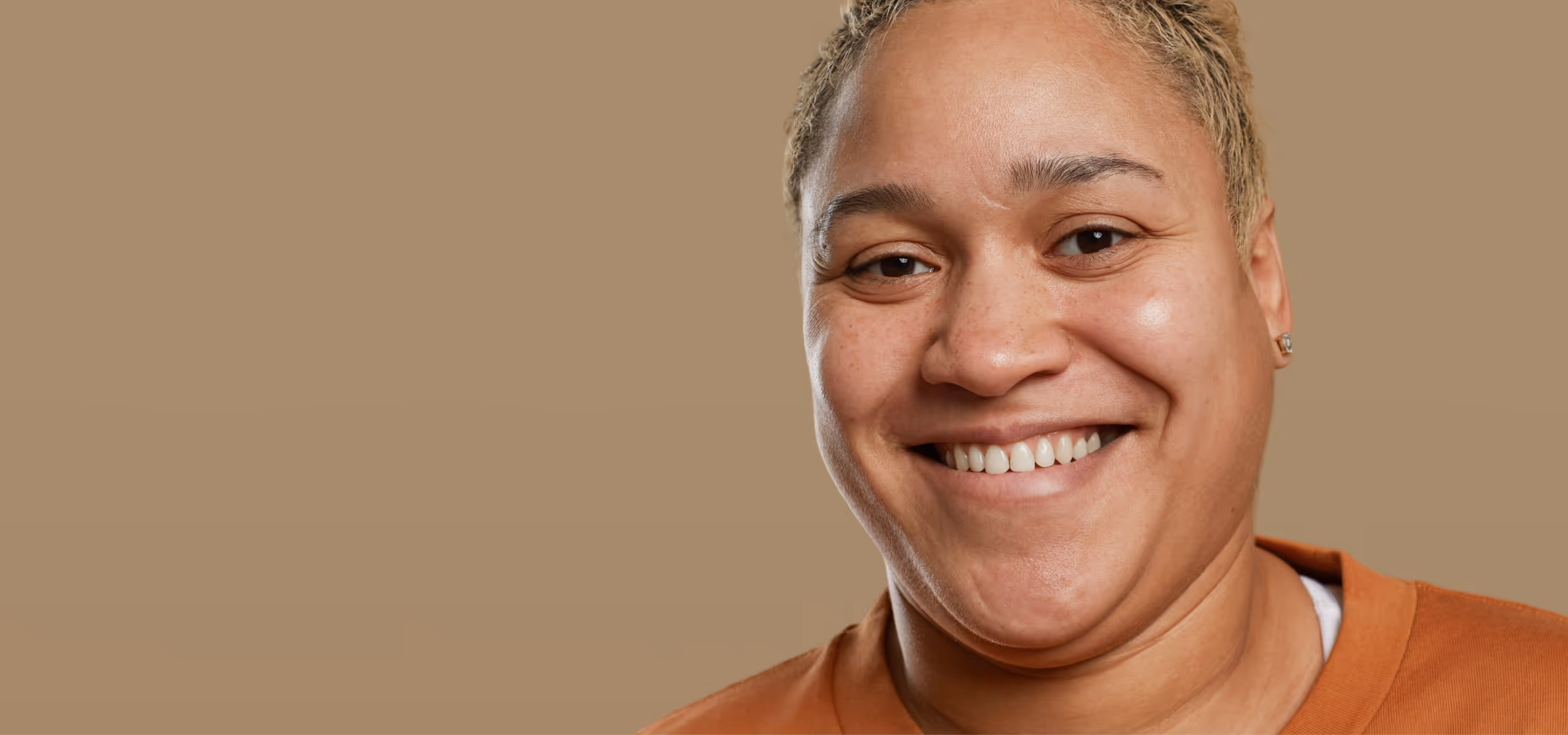 Smiling person with short blonde hair wearing an orange shirt against a beige background.