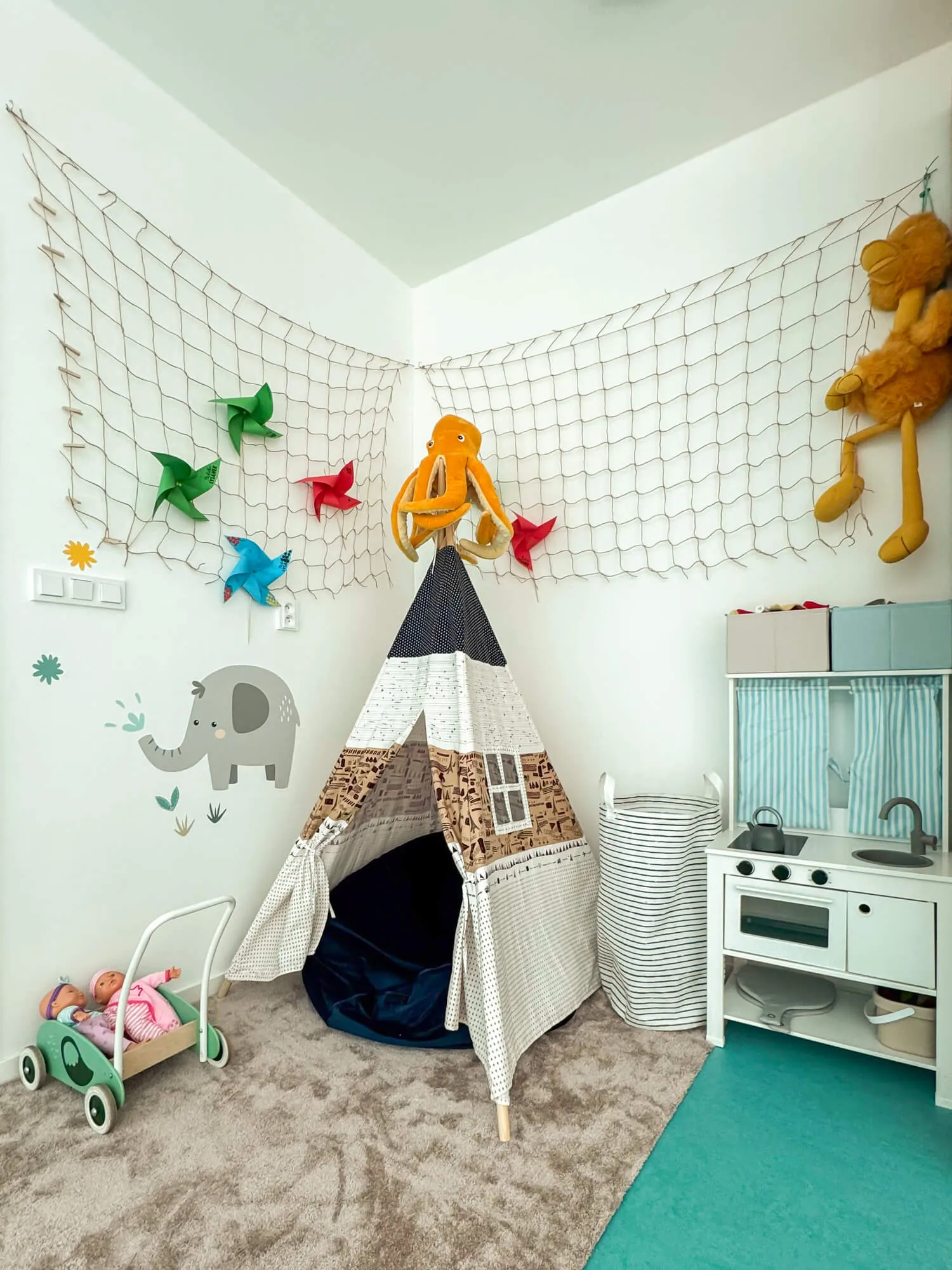 Dětský pokoj se šedým kobercem, teepee stanem, kuchyňkou a hračkami na podlaze.