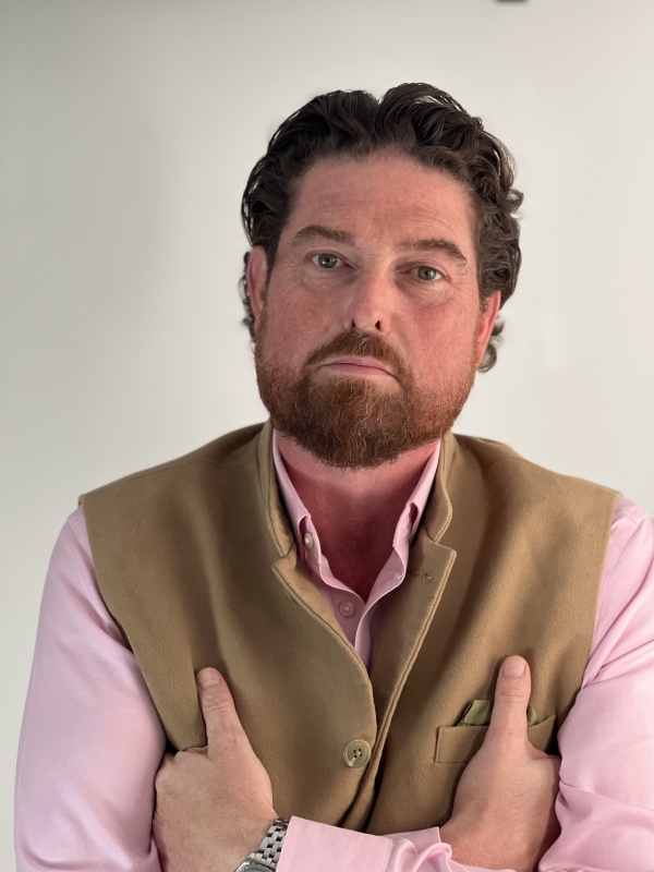 Image of Bruce wearing a tan vest over a light pink shirt, arms crossed, against a plain light background.