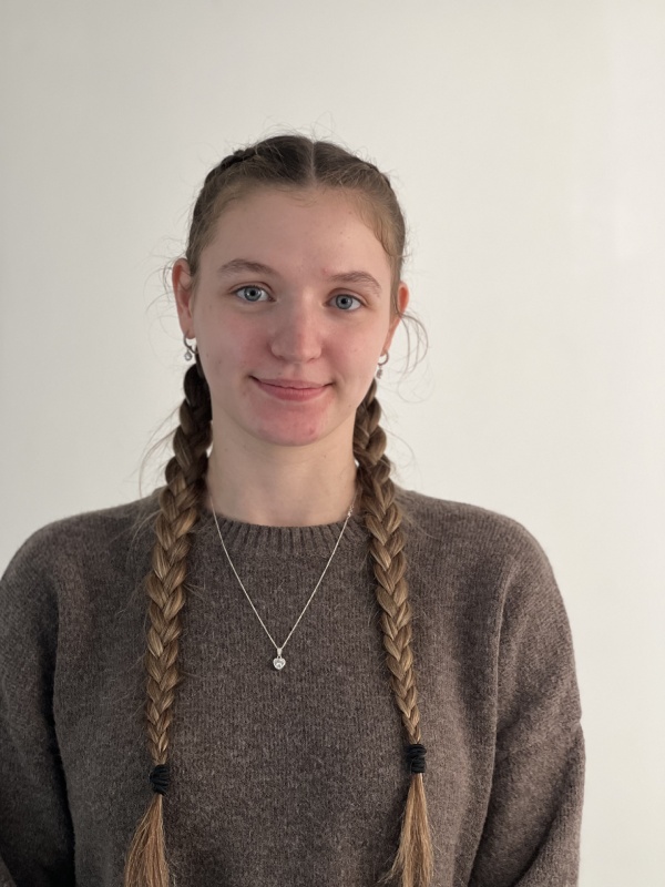 Young woman with long blonde hair styled in two braids wearing a brown sweater and a silver necklace with a round pendant, smiling gently against a plain light background.