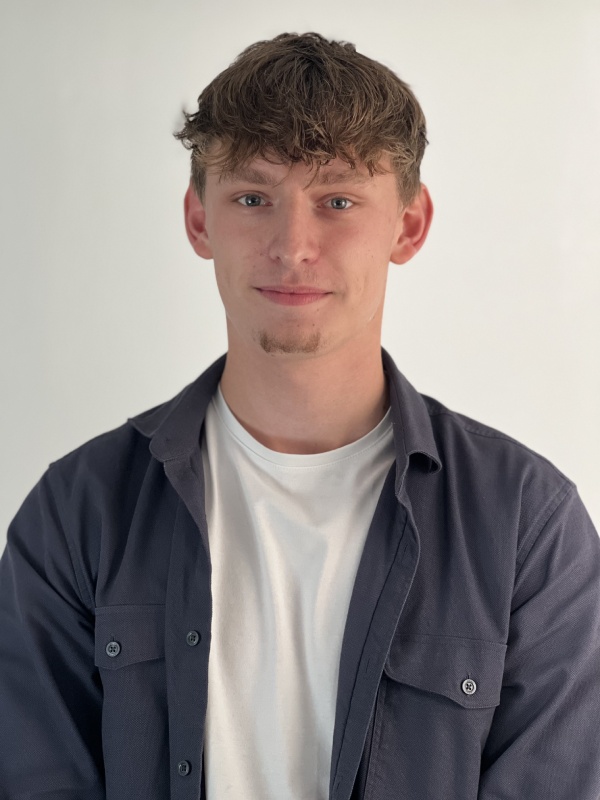 Young man with short brown curly hair and light facial hair, wearing a white t-shirt under an unbuttoned dark gray shirt, standing against a plain light background.