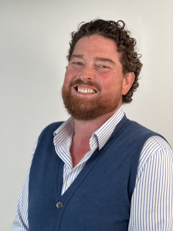 Smiling man (Bruce) with curly brown hair and beard wearing a blue vest over a white shirt with blue stripes against a plain light background.